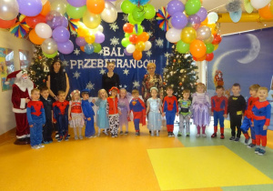 Grupa dzieci stoi z panią dyrektor Marią Królikowską, p. Anią i p. Natalią na tle dekoracji, pomiędzy przystrojonymi choinkami.